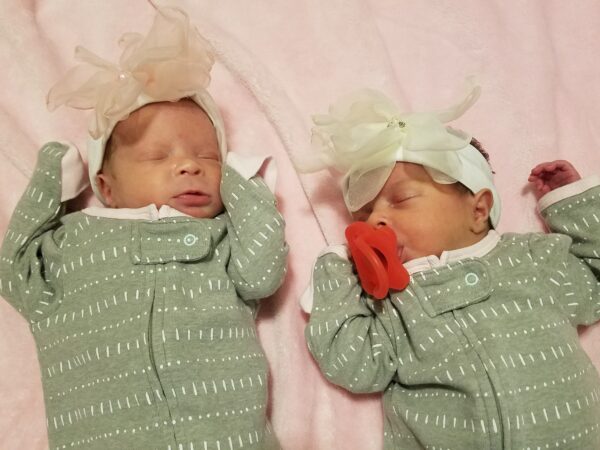 Newborn twins in green pajamas with bows in their hair sleeping on their back.