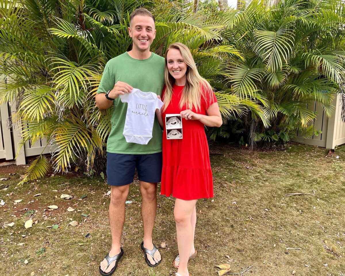 Couple standing in front of palm trees outside holding a baby onesie announcing a pregnancy. 