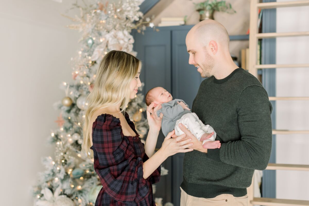 Young couple holding a newborn and looking at the baby in front of a Christmas tree. 