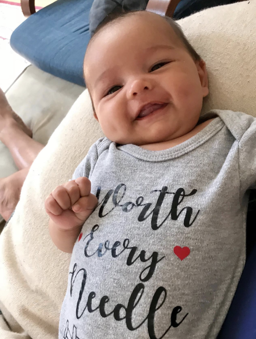 Infant smiling at the camera wearing a "onesie" that says, "Worth Every Needle"