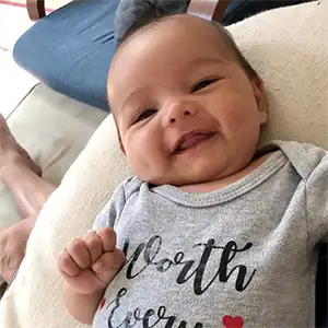 Infant smiling at the camera wearing a "onesie" that says, "Worth Every Needle"